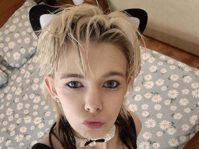 Close-up photo of LaurenceCloud, blonde hair, blue eyes, wearing a black and white outfit with a bow tie, looking up, daisy-patterned bed in background, slight messiness, intense gaze, sexual tension.