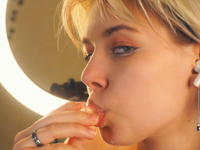 Close-up of LaurenceCloud with short blonde hair, blue eyes, and fair skin, sucking on their fingers with a ring on the right hand. Warm lighting highlights their sensual expression.
