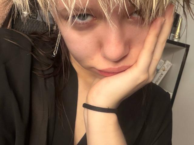 Foto em close-up de LaurenceCloud, uma mulher loira com cabelo molhado, vestindo roupa preta e usando uma pulseira preta, com a mão apoiada em... Bochechas rosadas, olhos azuis que fitam de forma sedutora, lábios levemente entreabertos. Muito atraente..