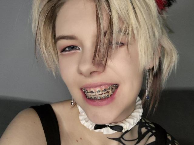 Close-up photo of LaurenceCloud, blonde with dark streaks, smiling with metal-studded teeth, wearing a black strap top and white choker with black bow, gray background, intense sexual tension.