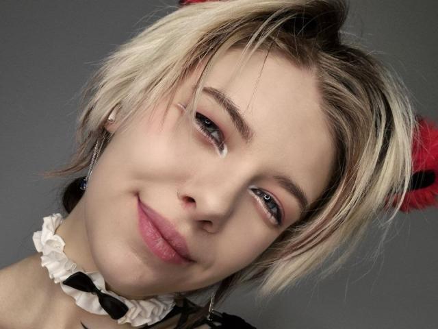 Close-up de LaurenceCloud, uma jovem com cabelo loiro curto e desalinhado, olhos azuis e um sorriso discreto. Ela está usando um colar branco com babados, com um fundo cinza. Muito fofo.