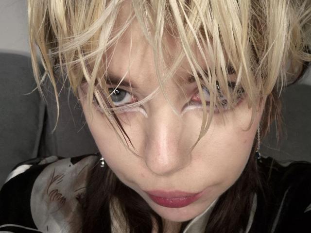 Foto em close-up de LaurenceCloud, com cabelo loiro e desalinhado cobrindo um olho, olhar azul intenso, batom vermelho, sorriso discreto. jaqueta preta, forte tensão sexual, fios de cabelo parcialmente cobrindo o rosto, fundo sombrio..