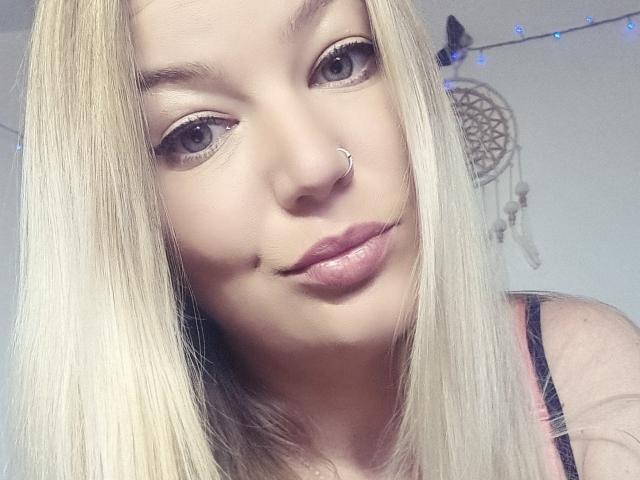 Selfie de perto de AlmaDeAngel69, cabelo loiro, piercing no nariz, pele clara, sorriso discreto, top com alças pretas, sonho apanhado (dreamcatcher) em fundo, luzes de fada azuis, leve tensão sexual, iluminação suave, pose casual.