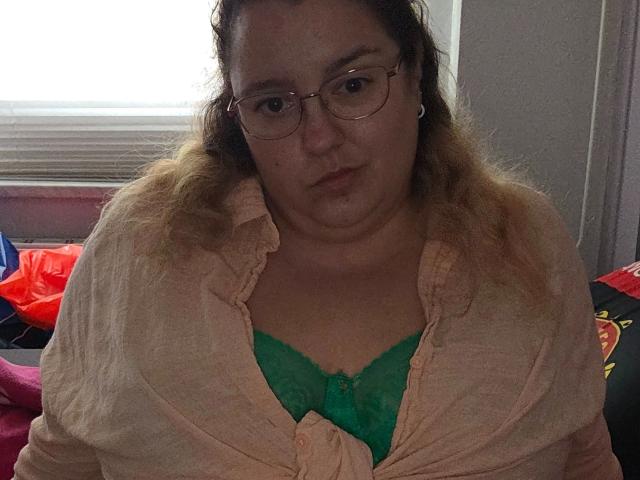 Fotografia di una donna formosa, Sarah-kommt-hot, con gli occhiali, che indossa una camicia color pesca aperta che rivela un reggiseno di pizzo verde. Lei guarda direttamente la telecamera, con un'espressione leggermente infastidita. Una finestra luminosa dietro di lei.