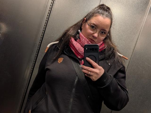 Fotografia de Sarah-kommt-hot com cabelo castanho preso em um rabo de cavalo, usando óculos, casaco preto, cachecol vermelho, segurando uma bolsa preta. telefone, tirando uma selfie em um elevador cinza com textura. Muito quente.