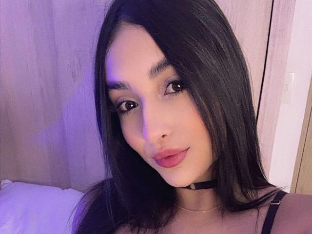 Selfie de perto de EmmaWaltter, uma jovem com cabelos longos e pretos, usando um colar preto e um top sem alças. Seus lábios estão ligeiramente entreabertos, e ela olha diretamente para a câmera com uma expressão sedutora..