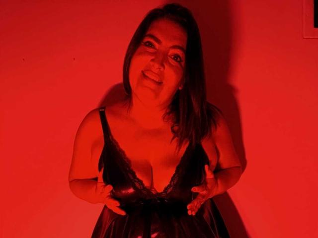 Photo éclairée en rouge de Lina69, avec une silhouette corpulente, souriante, penchée en avant, tirant sur de la lingerie en dentelle noire pour révéler une grande partie de son corps. Seins. Elle tient le tissu avec les deux mains, adoptant une pose séduisante..