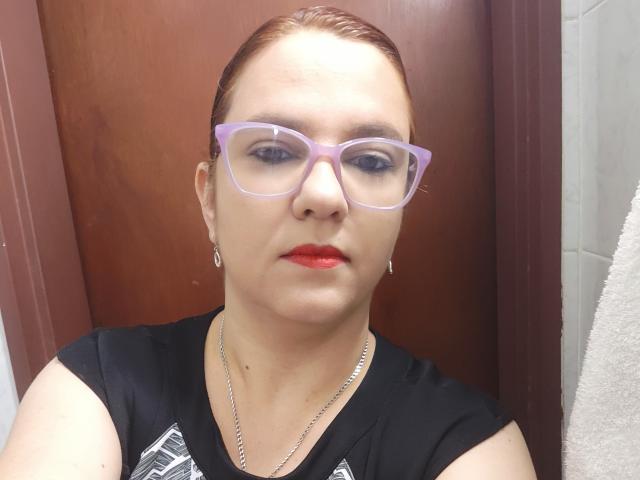 Amelia Wilson tiene el cabello castaño peinado hacia atrás, usa gafas de montura morada, lápiz labial rojo y una camiseta negra sin mangas. Logotipo blanco. Ella está mirando directamente a la cámara, transmitiendo una vibra segura y atractiva..