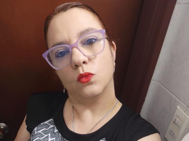 Amelia Wilson ha la pelle chiara, rossetto rosso e indossa occhiali viola, una maglietta nera con una grafica bianca e una collana d'argento. La sua espressione è leggermente corrucciata, creando un'aria sexy e sicura di sé..