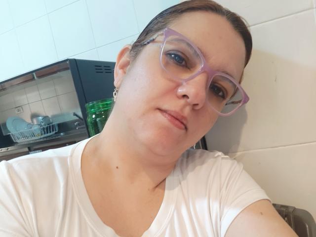Amelia Wilson, de piel clara, con cabello castaño recogido en una coleta, usando gafas moradas y una camisa blanca, apoyada contra una pared, lavando los platos al fondo. Un encanto adorable y un aire intelectual..