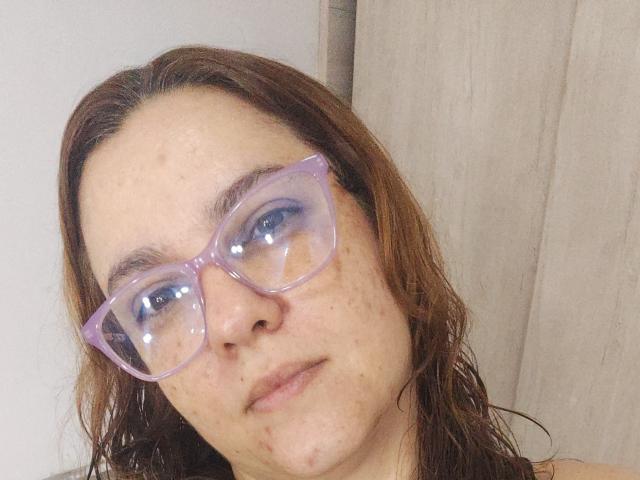 Selfie de perto de AmeliaWilson, com cabelo castanho molhado, usando óculos grandes, roxos e refletivos. Ela tem a pele clara com sardas, e parece um pouco deslocada da câmera, com uma expressão neutra. O fundo é uma cortina bege..