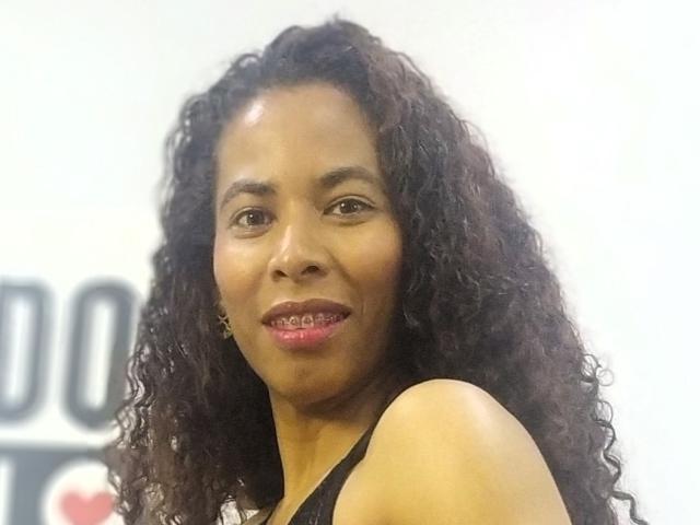 Fotografia em close-up de RebecaDuval, com cabelo cacheado escuro, olhos castanhos e batom rosa. Ela está sorrindo, mostrando levemente os dentes. Ela está vestindo uma blusa preta, com um fundo branco. Muito atraente..