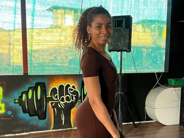 Rebeca Duval steht mit leicht abgewandtem Rücken und lächelt in die Kamera. Sie trägt ein eng anliegendes schwarzes Kleid, das ihre weiblichen Formen betont. Der Hintergrund zeigt farbenfrohe Graffiti und einen weißen Lautsprecher..