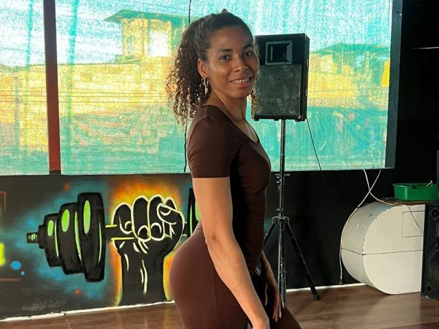 Rebeca Duval está de frente, virada para a esquerda, sorrindo, com cabelos cacheados, vestindo um vestido marrom justo que realça suas curvas. seios grandes. Uma tela de projetor com grafites coloridos e um alto-falante ao fundo..