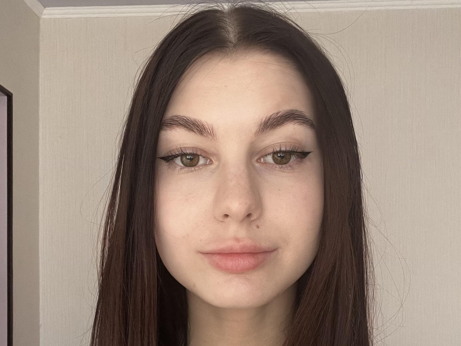 DeonnaDuriga has pale skin, green eyes with black eyeliner, brown eyebrows, and long brown hair. She's looking at the camera with a slight smile, showing her pink lips.