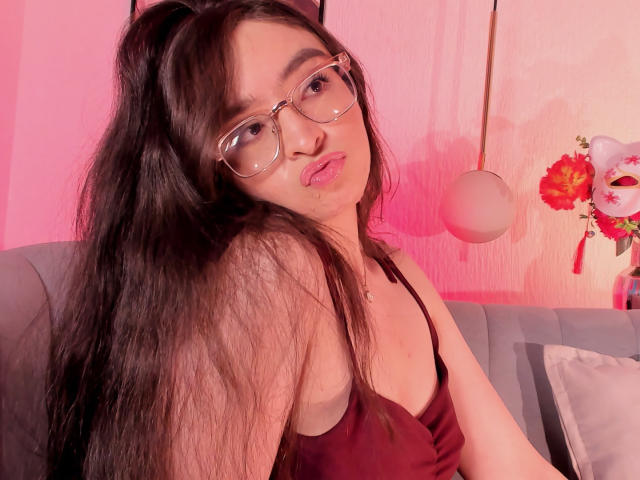 EvaMcAllister, avec ses longs cheveux noirs, portant des lunettes et un débardeur noir, regardant par-dessus son épaule avec une expression légèrement ouverte. bouche. Éclairage rose et rouge, fleurs floues en arrière-plan.