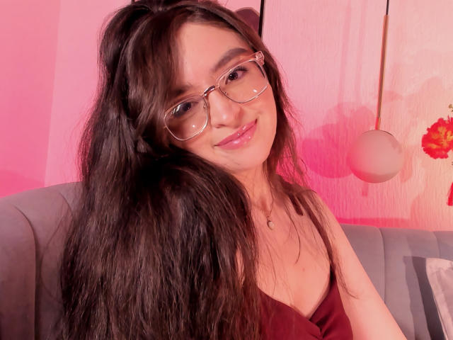 EvaMcAllister, una chica atractiva con cabello negro largo y gafas, está sentada en un sofá, vistiendo una blusa negra con escote pronunciado, iluminada con luz rosa. fondo, sonrisa suave, pose relajada, tensión sexual, ambiente sensual..