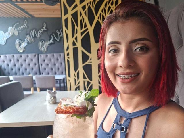 Fotografie einer rothaarigen Frau mit heller Haut, die lächelnd in die Kamera schaut und ein blaues Top mit Halter trägt. Sie hält ein Dessert. mit roter Soße und einem grünen Blatt. Restaurant im Hintergrund mit geometrischer Dekoration. Verdammt süß..