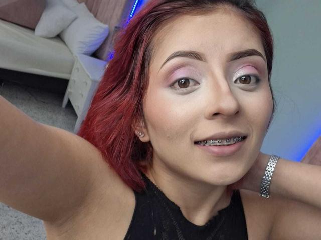 Close-up selfie of LizBonnet with red hair, light skin, and purple eyeshadow. She's smiling with a silver lip piercing, wearing a black top and a silver bracelet. Living room background.