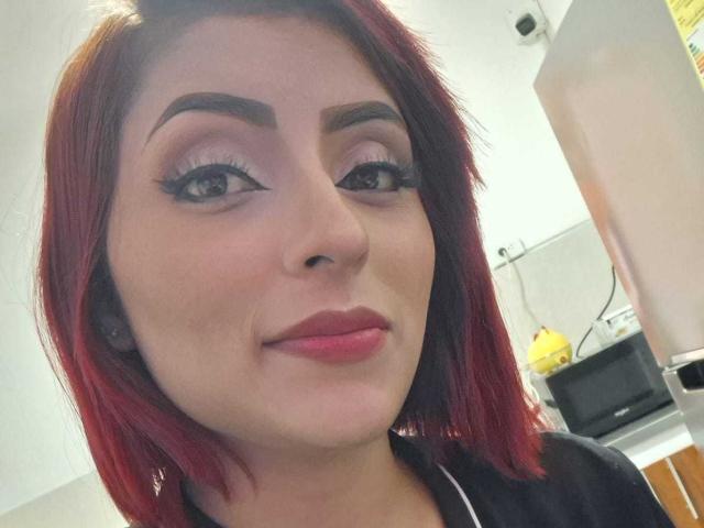 Autofoto de primer plano de LizBonnet, una mujer pelirroja con cejas pobladas, delineador de ojos negro y lápiz labial rosa. Está sonriendo ligeramente, mostrando un poco de escote, y lleva una blusa negra. Iluminación interior brillante.