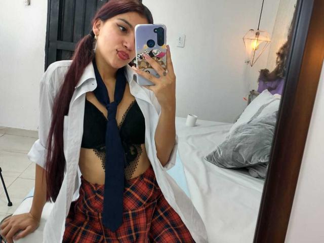 Noemi Ferrer, incroyablement sexy, prend un selfie devant un miroir. Elle porte une chemise blanche ouverte, un soutien-gorge en dentelle noire et une jupe à carreaux rouges, avec une cravate noire qui pend lâchement. Humectant ses lèvres de manière séduisante.