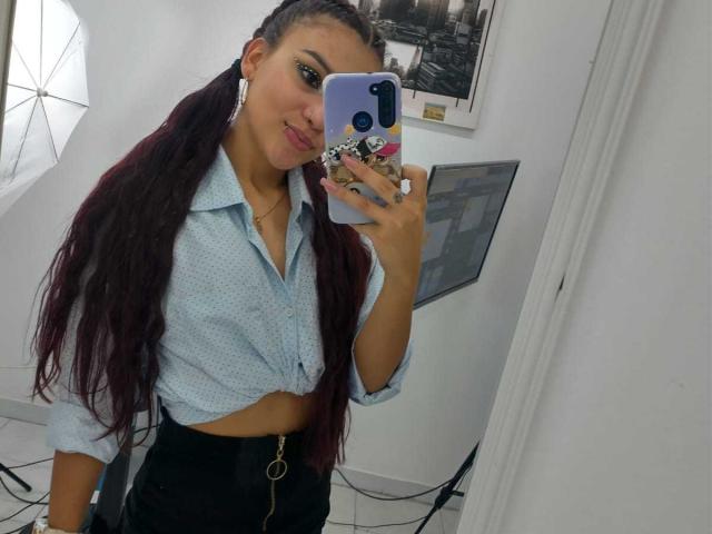 Noemi Ferrer prenant un selfie avec un smartphone, portant une chemise bleu clair nouée et un pantalon noir, avec de longs cheveux foncés. tenant le téléphone dans la main droite, tension sexuelle subtile, pose décontractée..
