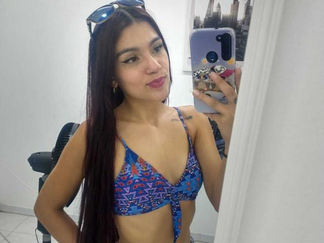 Noemi Ferrer se prend en photo dans un miroir, portant un haut de bikini bleu à motifs, avec de longs cheveux noirs, un anneau au nez, et lunettes de soleil sur sa tête. Elle tient un téléphone avec une coque colorée..