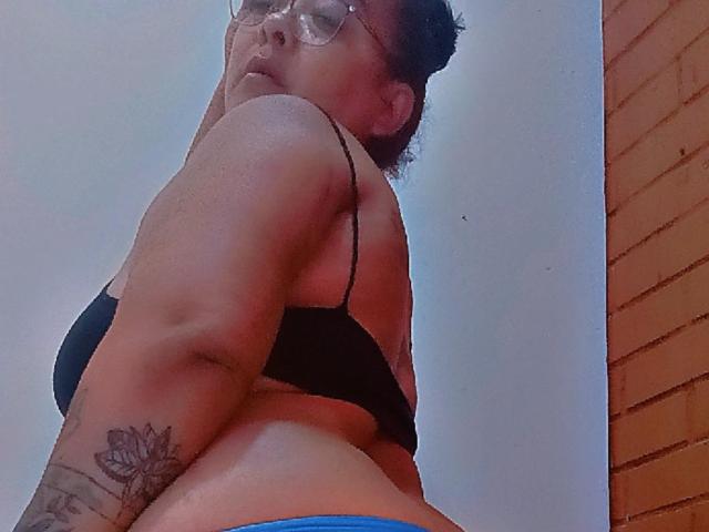 Fotografia tirada de baixo, mostrando SussanHott, usando óculos e um sutiã preto, olhando por cima do ombro. Tatuagem visível no braço superior, calcinha azul, ligeiramente desfocada. O fundo inclui uma parede de tijolos. Incrivelmente sexy..