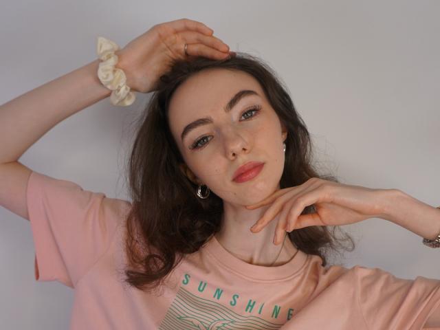 Shari Ravetto, avec sa peau claire et ses cheveux brun foncé, porte une chemise rose avec l'inscription "SOLEIL". Les mains levées, une sur la tête, l'autre près du menton. Un élastique blanc au poignet. Tension sexuelle, regard séduisant.