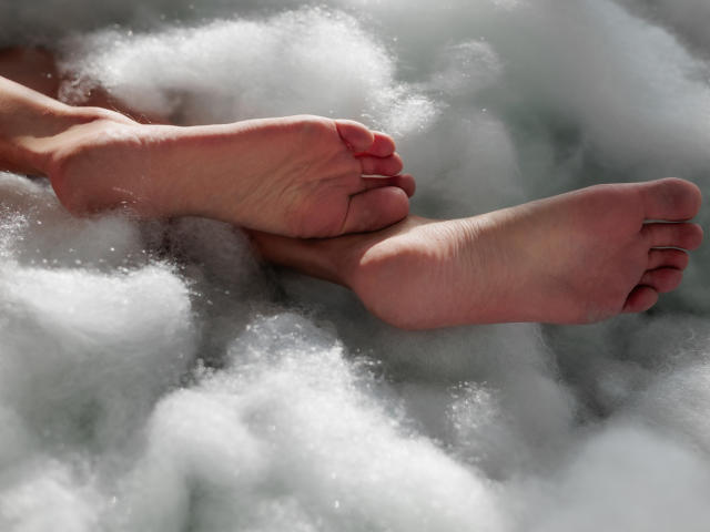 Primer plano de los pies descalzos de MiaCuteAngel, con las plantas hacia arriba, con uñas de los pies rojas, rodeados de nubes blancas brillantes y esponjosas. Los pies muestran un ligero tono rosado en la piel, los dedos relajados. Ligera tensión erótica..