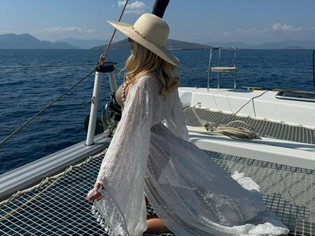 AvaMadyson est agenouillée sur le pont d'un voilier, vêtue d'une robe blanche en dentelle et d'un chapeau à larges bords, le dos tourné vers la caméra. Océan et montagnes en arrière-plan, ciel bleu au-dessus. Lumière du soleil, tension sexuelle..