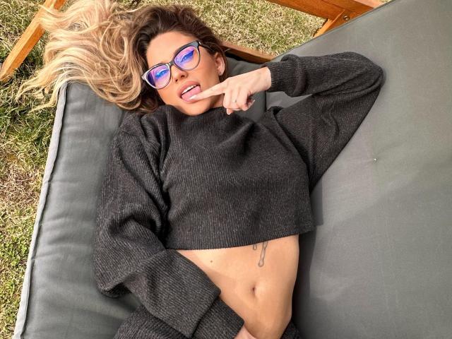 AvaMadyson lies on a gray couch, wearing blue sunglasses and a black crop top, exposing her midriff and small tattoo. She points a finger while smiling. Blonde hair spreads out.