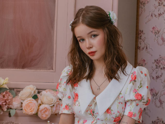 ViolettaFane, met golvend bruin haar, draagt een witte blouse met bloemen en een grote kraag, en een groene haarspeld. Ze kijkt recht in de camera, haar lippen zijn lichtjes open. In de achtergrond staan roze rozen..