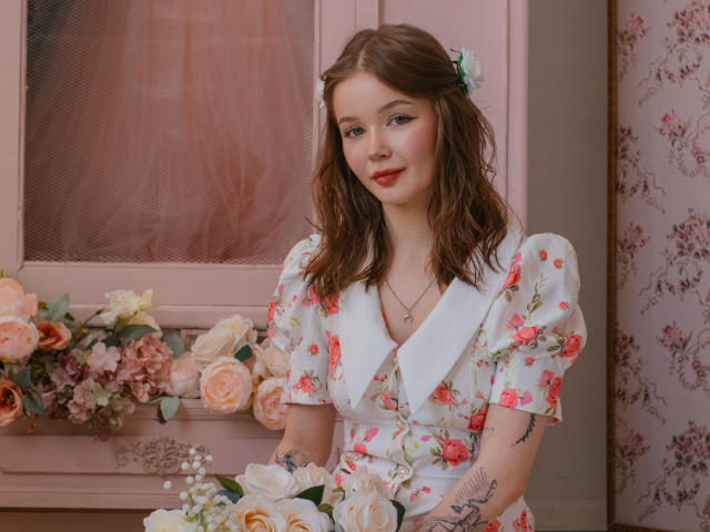 Violetta Fane mit welligem, braunem Haar, einem Blumenkleid und einem weißen Kragen, die Rosen hält. Rosa, blumige Tapete und ein Fenster im Hintergrund. Sie schaut direkt in die Kamera, mit einem leichten Lächeln, und hat sichtbare Tattoos am Arm..