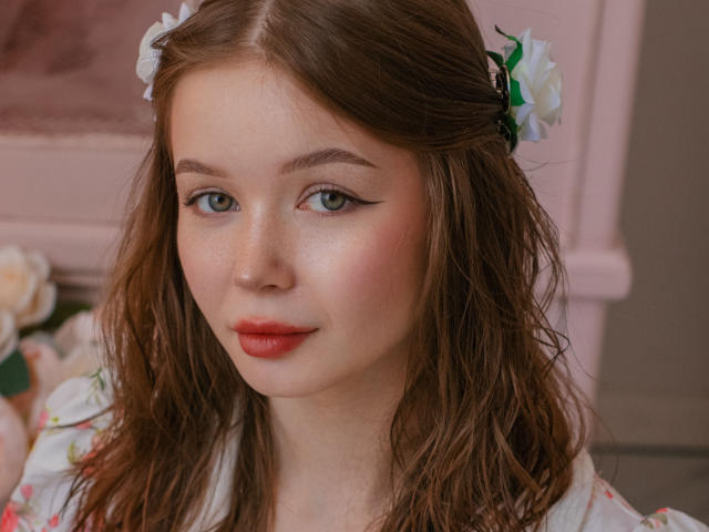 Close-up photo of ViolettaFane with fair skin, blue eyes, and red lipstick. She has brown hair with white flowers, wearing a floral dress. Background has blurred flowers and pink furniture. Fucking gorgeous.