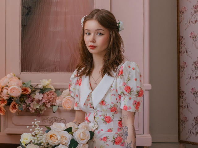 ViolettaFane è in una stanza rosa, e tiene in mano un bouquet di rose. Indossa un abito bianco con fiori, con rose rosse, ha i capelli castani bagnati e dei tatuaggi sul braccio sinistro. Assolutamente stupendo..