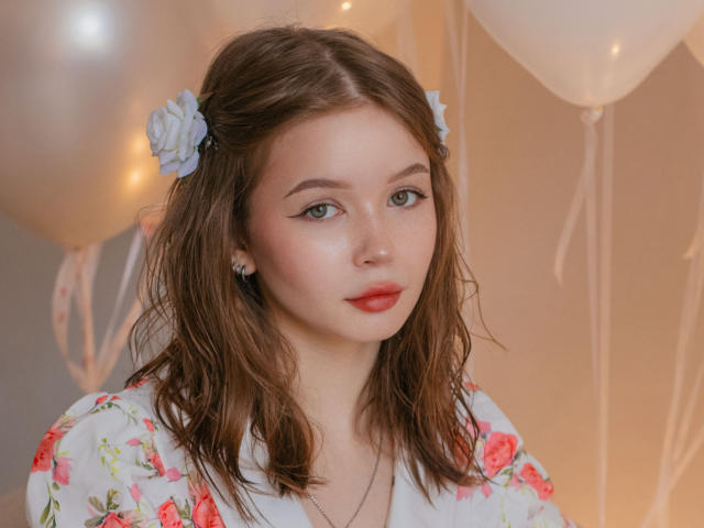 Fotografia de Violetta Fane, uma jovem com pele clara, olhos azuis e batom vermelho, vestindo um vestido branco com estampas florais. Presilha de cabelo em formato de flor azul. Balões desfocados ao fundo. Absolutamente lindos..
