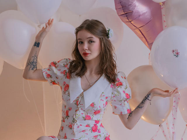 ViolettaFane, avec des cheveux bruns mi-longs, une robe à fleurs, un col blanc, tenant des ballons blancs, des tatouages sur les bras, et une expression douce. Décoration de fond avec des ballons roses et blancs, tension sexuelle subtile, expression confiante, pose ludique..