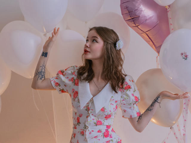 ViolettaFane, con cabello largo y castaño, vestida con un vestido blanco con estampado floral y cuello, rodeada de blanco y rosa. globos. Ella está sosteniendo un globo con su mano izquierda, y se pueden ver tatuajes en sus brazos..