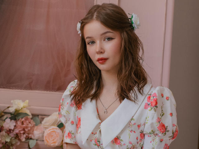 Violetta Fane, con i capelli castano ondulati e un abito bianco con fiori, è in piedi di fronte a una finestra rosa. Indossa delle forcine con i fiori, una collana e ha il rossetto rosso. Composizione floreale sullo sfondo.