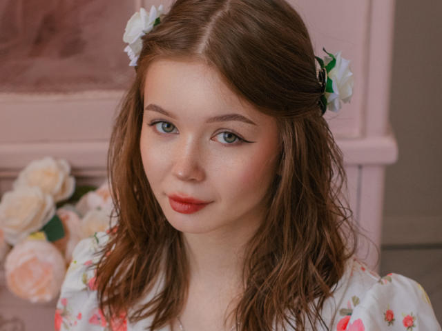 Close-up foto van ViolettaFane, met een lichte huid en blauwe ogen, die een witte jurk met bloemenmotief draagt, rode lippenstift en wit bloemen in haar bruine haar. Zachte roze tint, lichte glimlach, bloemenachtergrond, seksuele spanning.