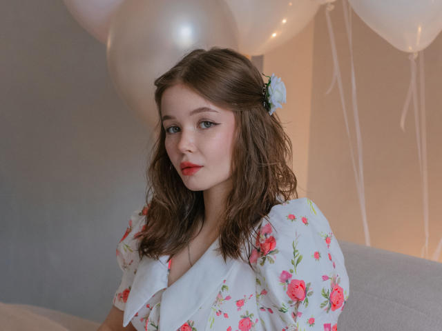 Fotografia di ViolettaFane, una donna dalla pelle chiara con capelli castani ondulati, che indossa una camicetta bianca con fiori e un fiore blu. nei suoi capelli. Palloncini bianchi sullo sfondo. Sta guardando la fotocamera con un leggero sorriso, rossetto rosso e uno sguardo sottile e seducente..