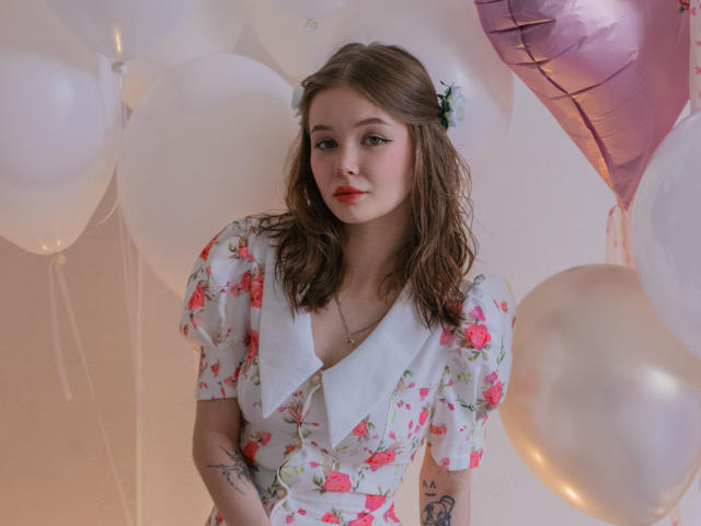 ViolettaFane está de pie en medio de globos blancos y rosas, vistiendo un vestido floral con un cuello blanco. Ella tiene lápiz labial rojo, cabello castaño ondulado y tatuajes visibles en su brazo. Qué lindo/a..