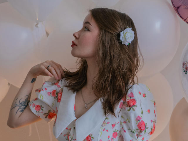 ViolettaFane, met bruin haar dat tot op haar schouders reikt, draagt een witte jurk met bloemen en heeft een bloem in haar haar. Ze kijkt naar de zijkant. Witte ballonnen vullen de achtergrond. Een tatoeage op haar linkeronderarm. Heel schattig.