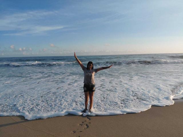 PauletteLoveX está de pie en una playa arenosa, con los brazos extendidos, mirando hacia el océano. Ella lleva una camiseta gris y pantalones cortos negros, con pequeñas huellas que conducen a sus pies. La luz del sol le daba en la espalda..