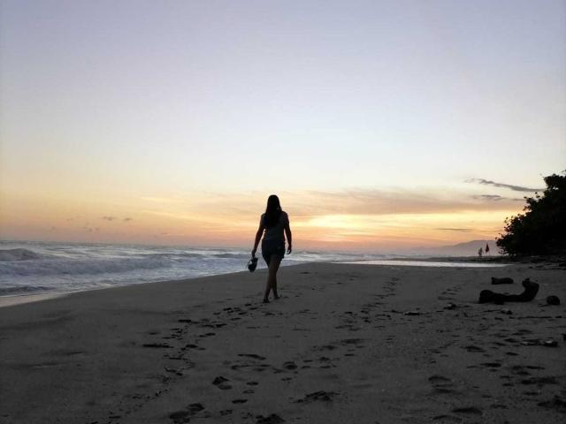 PauletteLoveX, con el pelo largo y silueteada, camina por una playa al atardecer, sosteniendo un teléfono en la mano derecha y vistiendo ropa oscura. vestido, olas en primer plano, degradado del cielo de naranja a azul.