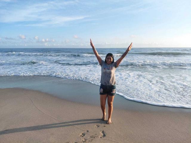 PauletteLoveX stands on a sandy beach with arms raised, wearing a gray shirt and dark shorts. Ocean waves crash in the background. She's barefoot with a shadow on the sand.