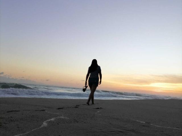 Silueta de PauletteLoveX caminando en una playa al atardecer, sosteniendo una cámara en la mano derecha, con olas en el fondo, y un degradado de color. cielo que va del naranja al azul, costa oscura, figura en sombra.