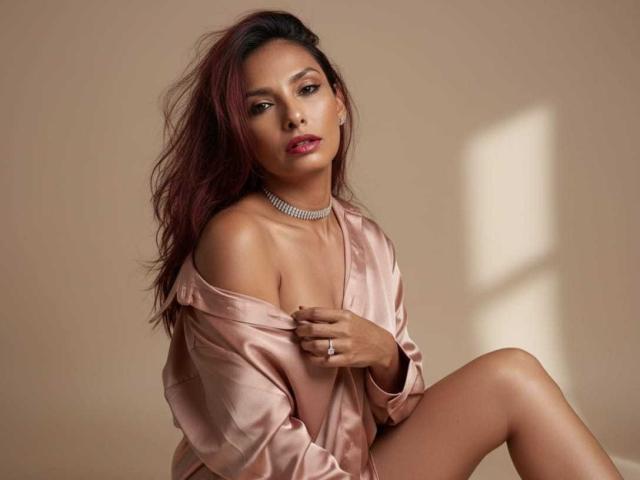 Photograph of RoxaneMoore with medium brown skin, red lipstick, and messy dark brown hair. She's wearing a silky pink off-shoulder shirt, choker necklace, and ring. She's sitting with one leg raised, looking seductively at the camera.