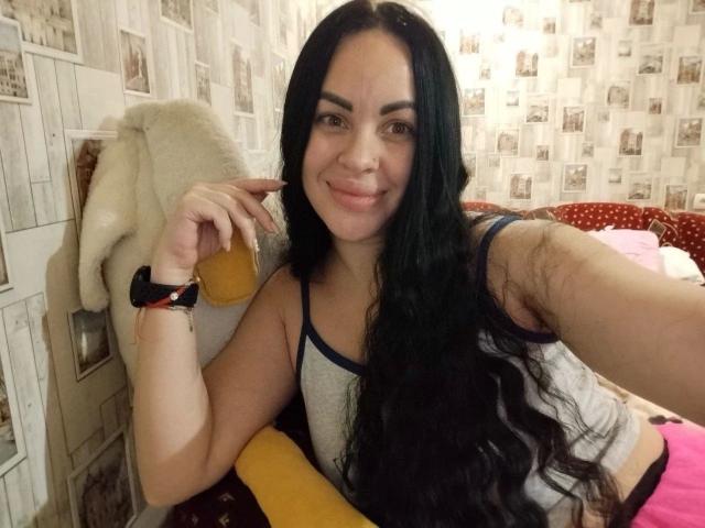 Eleonoracherry-hot, a hot chick with long black hair, smiling at the camera, wearing a grey tank top, black wristband, and pink shorts. Background includes patterned wallpaper and a stuffed animal.